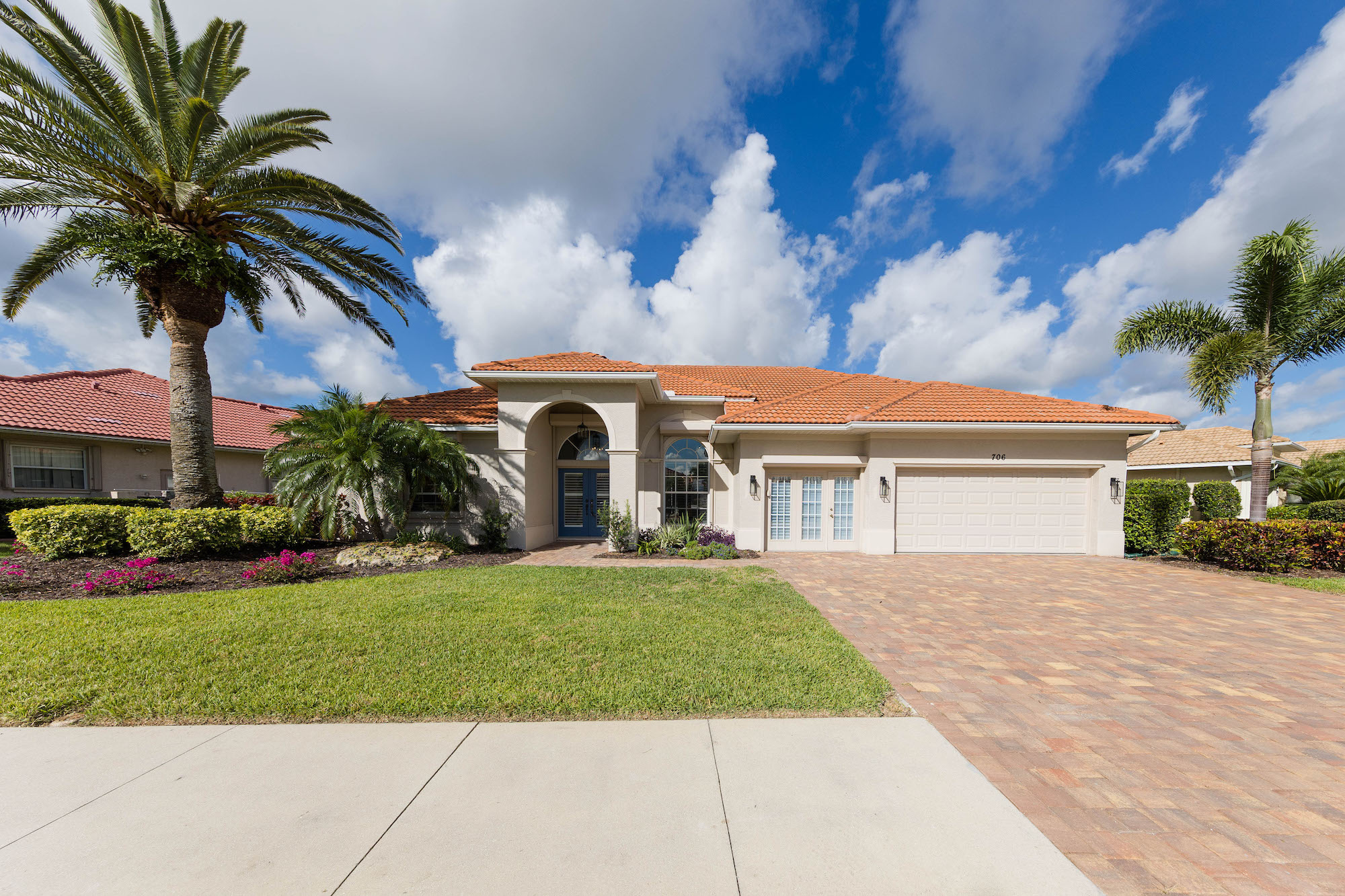 exterior of white home with orange tile roofing, brick driveway, palm trees and other flowers